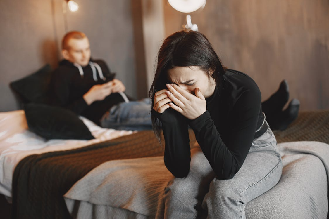 A woman wearing a black top and grey jeans crying while sitting on a bed