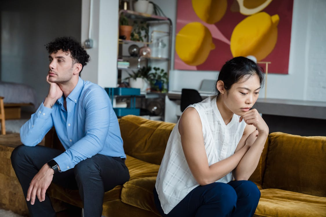 Distressed couple sitting apart on a mustard-colored sofa in a modern living room, looking away from each other.