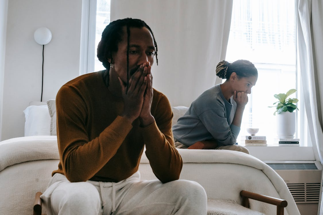Couple sitting apart in a bright bedroom, both appearing worried and deep in thought.
