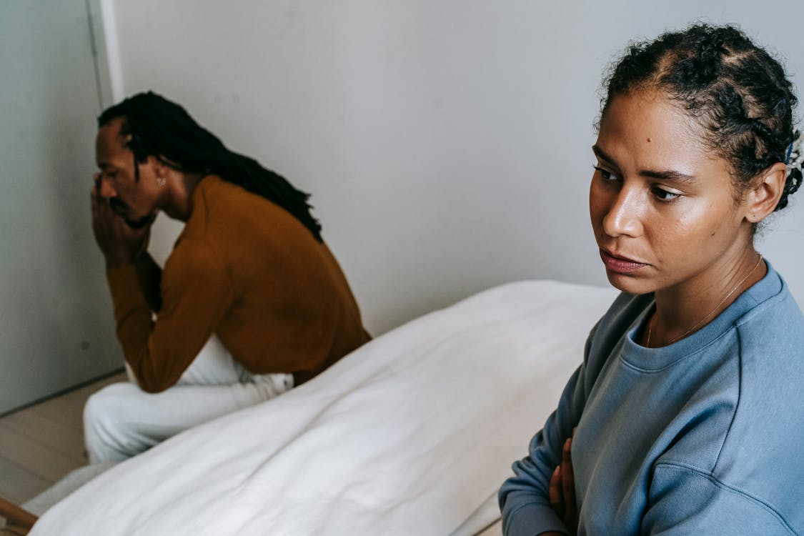 A sad man and woman, both seated on a bed, while the man has his hands near his face