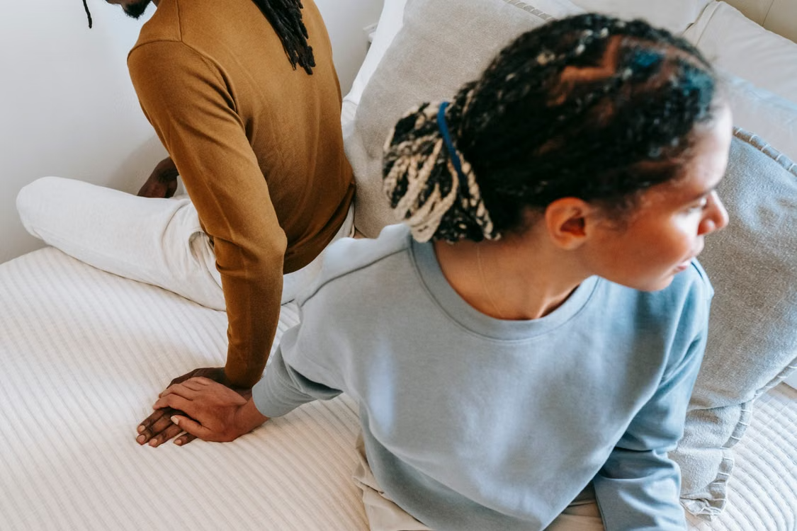 A couple sit on opposite sides of their bed facing away from each other, but both reach back to touch hands